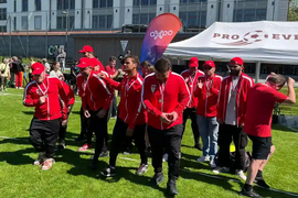 Eine Gruppe von Menschen in passenden roten Jacken und Hüten steht im Gras, einige klatschen und jubeln, vor einem weißen Veranstaltungszelt und einem bunten Oxpo-Banner, mit einem Gebäude im Hintergrund.