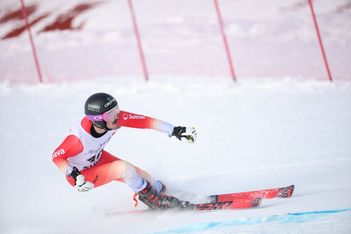 Ein Skifahrer in einem rot-weißen Anzug und mit schwarzem Helm macht während eines Rennens eine scharfe Kurve auf einer verschneiten Piste, im Hintergrund ist ein roter Zaun zu sehen.