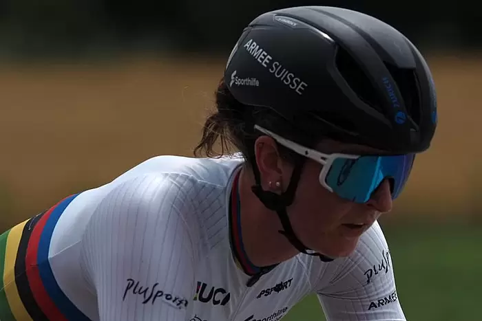 Ein Radfahrer mit schwarzem Helm, reflektierender Sonnenbrille und einem weißen Trikot mit Regenbogenstreifen und Logos fährt im Freien und konzentriert sich auf die Straße vor sich.