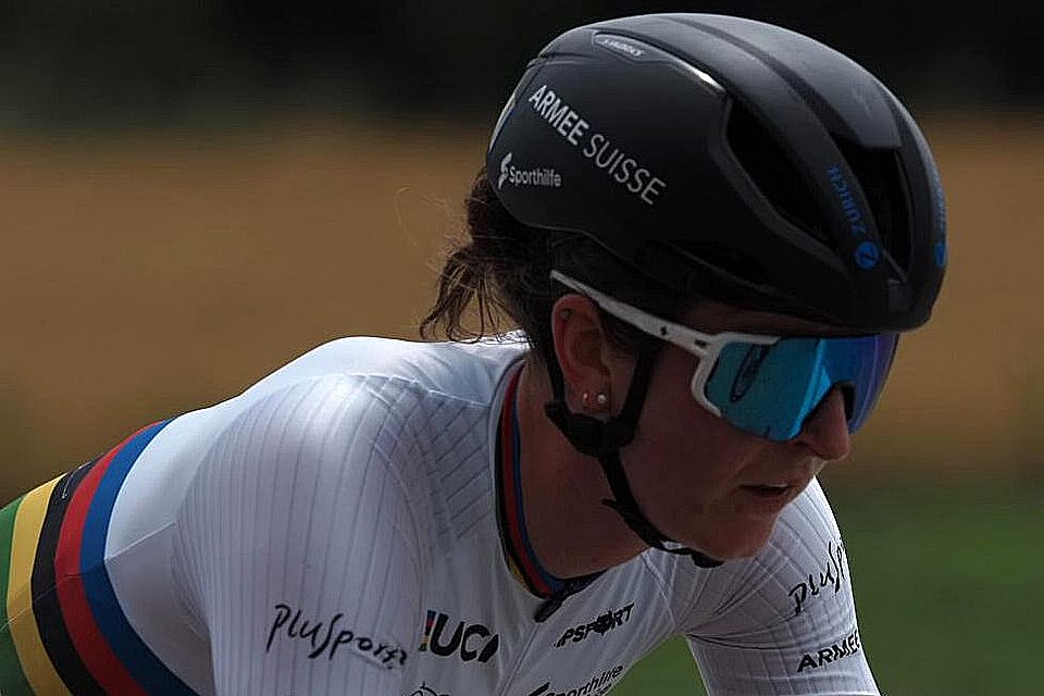 Ein Radfahrer mit schwarzem Helm, reflektierender Sonnenbrille und einem weißen Trikot mit Regenbogenstreifen und Logos fährt im Freien und konzentriert sich auf die Straße vor sich.