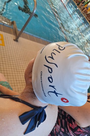 Ein Schwimmer, der eine weiße RivSport Oberemmental Badekappe trägt, sitzt mit dem Rücken zur Kamera am Rand eines Hallenbades, in dem im Hintergrund Menschen schwimmen.