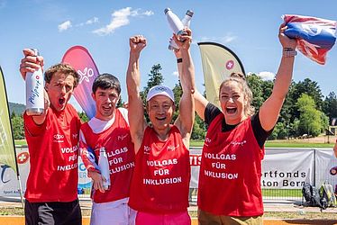 Vier lächelnde Sportler in roten Trikots stehen gemeinsam im Freien und recken jubelnd die Arme in die Höhe. Sie halten Jonglierkeulen und eine Fahne in den Händen, im Hintergrund sind bunte Banner und ein sonniger Himmel zu sehen.