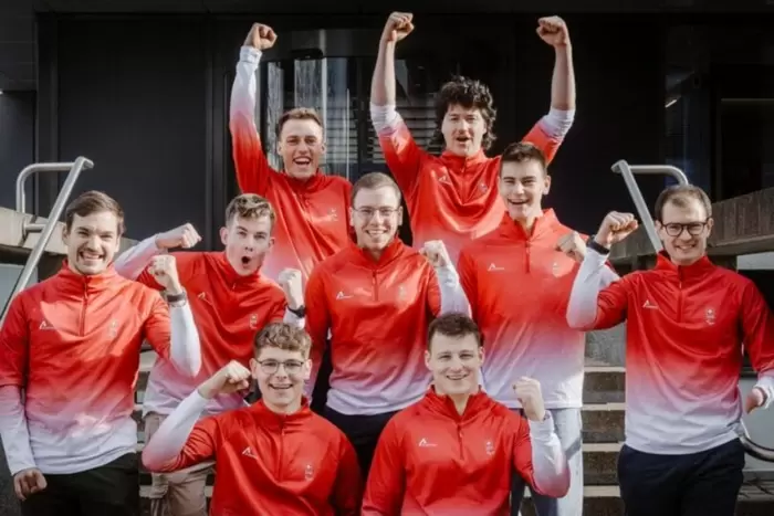 Neun junge Männer in farblich passenden rot-weißen Sportjacken stehen auf einer Treppe, lächeln und heben aufgeregt die Fäuste zum Feiern.