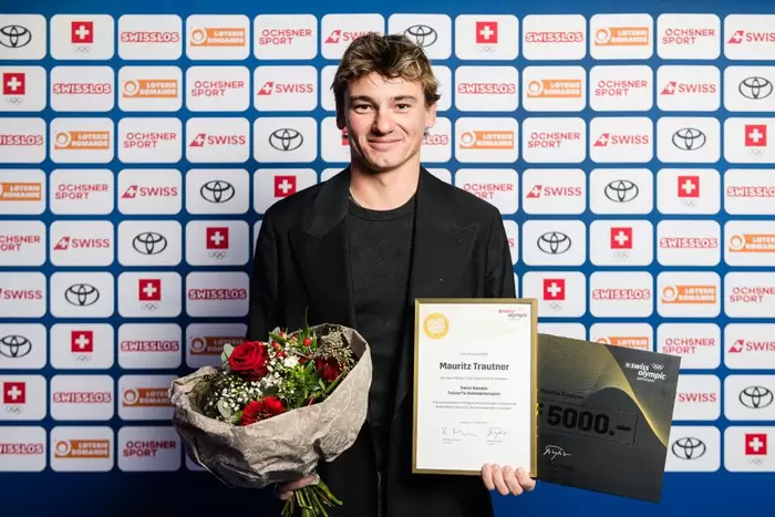 Mauritz Trautner steht lächelnd mit Blumenstrauss in der rechten und Award-Diplom in der linken Hand vor der Sponsorenwand von Swiss Olympic.