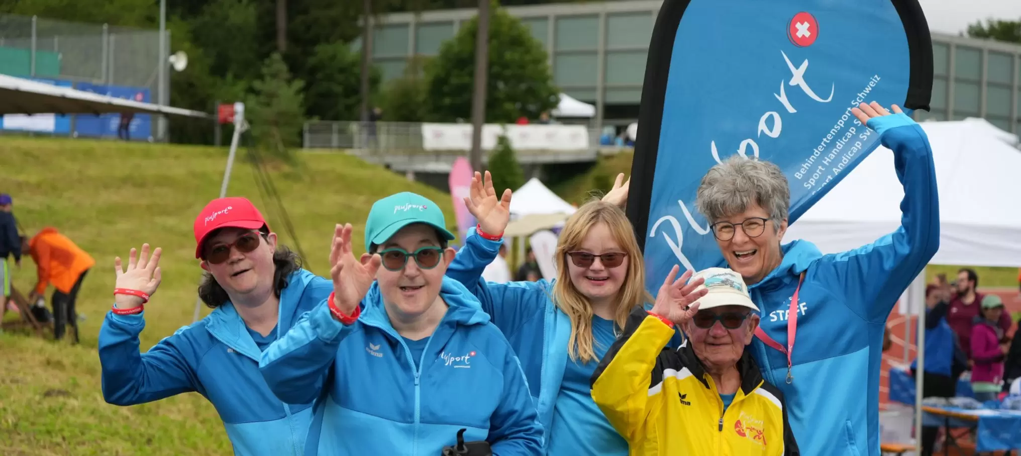 Fünf Personen in bunten Jacken und Hüten lächeln und winken während einer Veranstaltung im Freien in die Kamera. Sie stehen vor einem PlaySport-Banner, im Hintergrund sind Grasflächen und Zelte zu sehen.