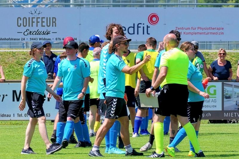 Eine Gruppe von Fußballspielern, Trainern und Schiedsrichtern in hellen Trikots steht auf einem sonnigen Fußballplatz und unterhält sich, während im Hintergrund am Zaun Werbung zu sehen ist.