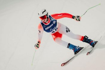 Ein Skifahrer in rot-weißem Anzug und Helm rast auf Schnee bergab, trägt eine blaue Allianz-Startnummer 45, hält Skistöcke in der Hand und lehnt sich in eine Kurve.