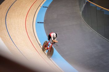 Ein Radfahrer, der einen Helm und eine Rennausrüstung trägt, fährt mit einem roten Fahrrad auf einer Indoor-Velodrombahn, wobei er sich in einen gebogenen Abschnitt mit blauen und hölzernen Fahrspuren hineinlehnt.
