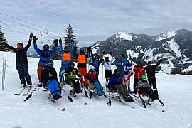 Eine Gruppe von Skifahrern, darunter auch Personen mit adaptiver Skiausrüstung, posiert gemeinsam auf einem verschneiten Berghang mit erhobenen Armen, umgeben von schneebedeckten Bäumen und Bergen im Hintergrund.