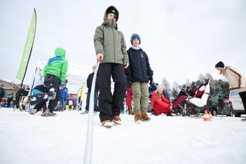Zwei winterlich gekleidete Kinder stehen im Schnee, eines hält einen weißen Stock. Im Hintergrund sind Menschen und Schlitten zu sehen, und an einem bewölkten Tag sind eine grüne Fahne und schneebedeckte Bäume zu sehen.