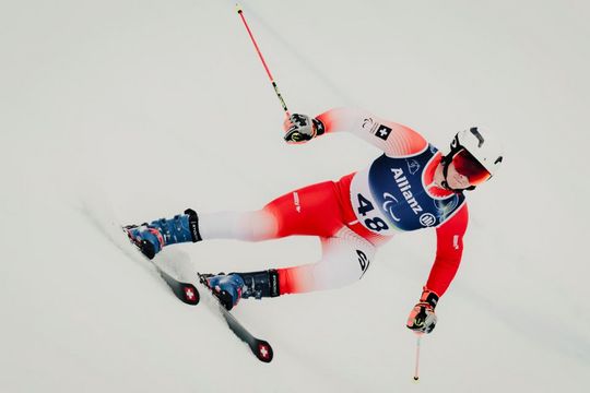 Ein Skifahrer in einem rot-weißen Anzug, mit Helm und Brille, fährt bei einem Slalomrennen bergab. Der Skifahrer lehnt sich auf einer verschneiten Piste in eine Kurve, hält Skistöcke in der Hand und trägt die Nummer 48 auf seiner Startnummer.
