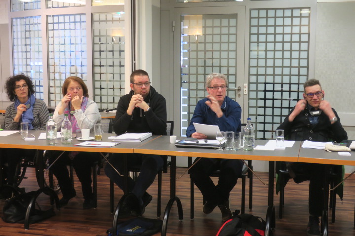 Fünf Personen sitzen an einem Konferenztisch in einem Besprechungsraum. Sie diskutieren. Auf dem Tisch liegen Papiere, Wasserflaschen und Gläser. Im Hintergrund ist eine Trennwand mit einem Gittermuster zu sehen.
