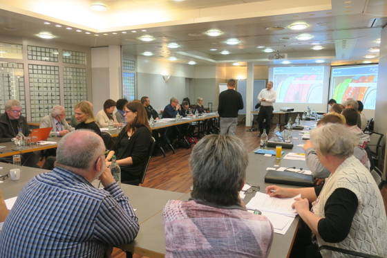 Eine Gruppe von Personen sitzt in einem Konferenzraum an rechteckigen Tischen. Zwei Vortragende stehen neben einem Bildschirm, auf dem eine bunte Folie läuft. Die Teilnehmer machen sich Notizen und verfolgen die Präsentation. Der Raum ist durch Deckenleuchten gut beleuchtet.