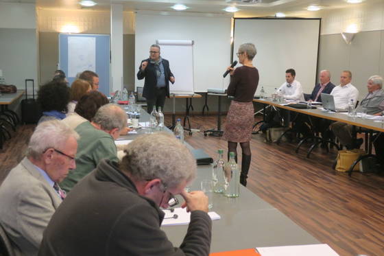 Eine Gruppe von Menschen sitzt in einem Konferenzraum an Tischen. Eine Frau steht mit einem Mikrofon da und blickt einem sprechenden Mann entgegen. Präsentationsbildschirme und Flipcharts sind zu sehen. Flaschen und Papiere liegen auf den Tischen.