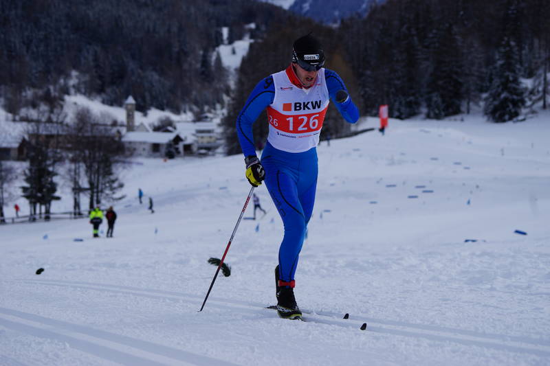 Ein Langläufer in blauem Anzug und schwarzer Mütze fährt auf einer verschneiten Loipe. Er benutzt Stöcke und trägt die Startnummer 126. Im Hintergrund sind schneebedeckte Bäume und Gebäude zu sehen.