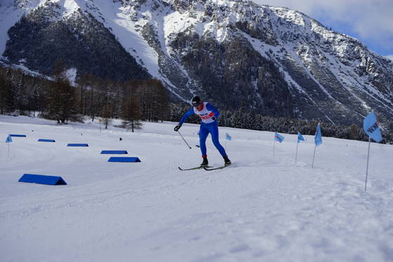 Eine Person in blauer Skiausrüstung läuft auf einer verschneiten Loipe mit Bergen im Hintergrund Skilanglauf. Blaue Fahnen säumen die Strecke unter einem klaren Himmel.