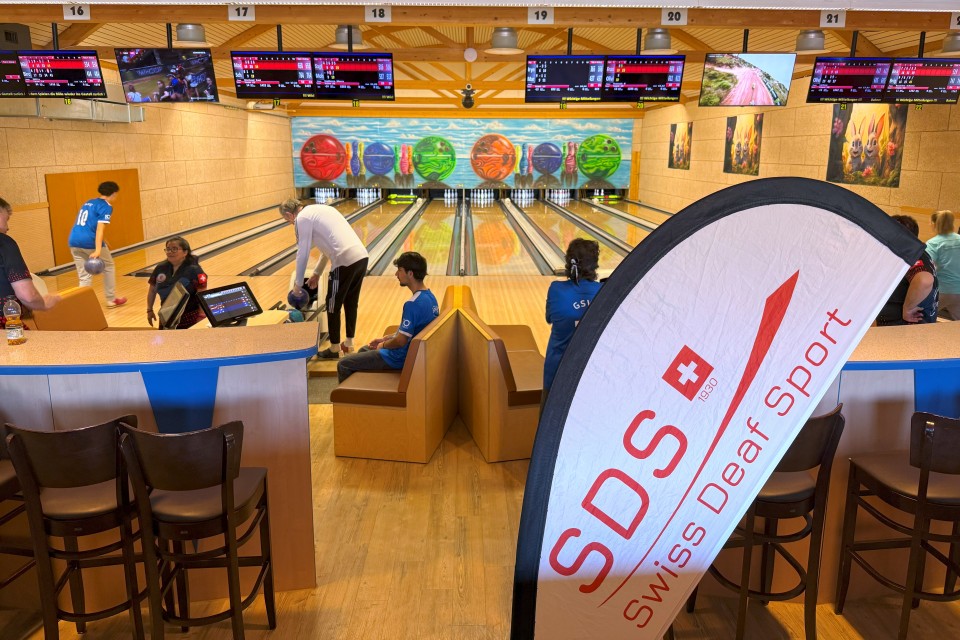 Eine Bowlingbahn mit Spielern und Zuschauern, darunter eine Gruppe in blauen Shirts. Im Vordergrund steht ein Banner mit der Aufschrift „SDS Swiss Deaf Sport“. Im Hintergrund sind Bowlingbahnen, Bildschirme und farbenfrohe Wandkunst zu sehen.