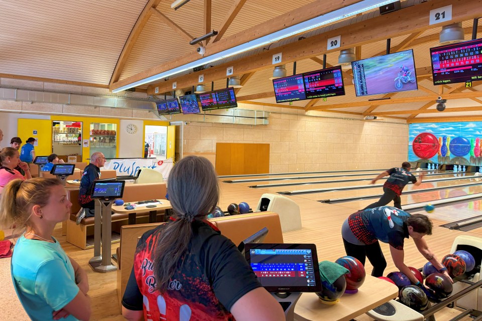 Eine Gruppe von Menschen in Bowlinghemden steht da und beobachtet, wie zwei Spieler mitten im Spiel in einer modernen Bowlingbahn bowlen und sich auf das Bowlen vorbereiten. Über den Bahnen werden auf digitalen Bildschirmen die Spielstände angezeigt.