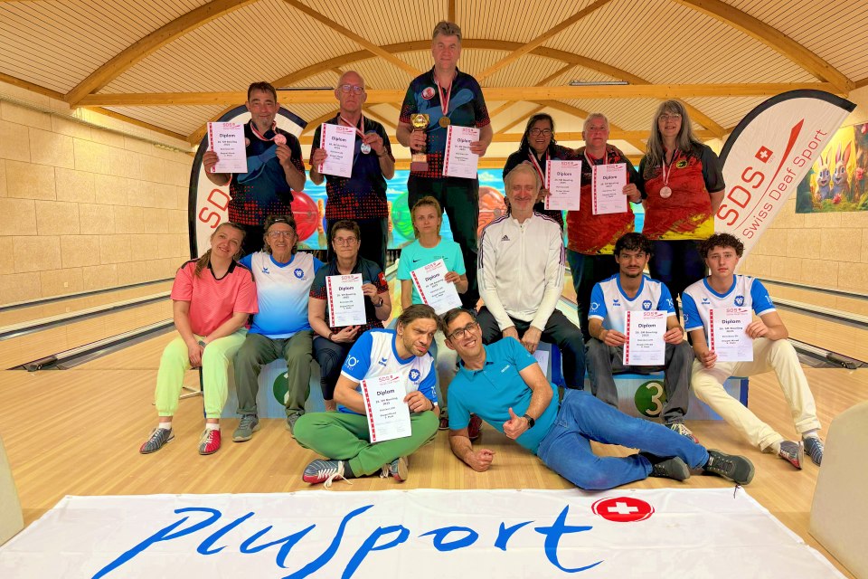 Eine Gruppe von Sportlern und Trainern posiert auf dem Podium einer Bowlingbahn und hält Urkunden und Medaillen in den Händen. Sie lächeln und tragen Sportuniformen, vor denen ein Plusport-Banner weht.