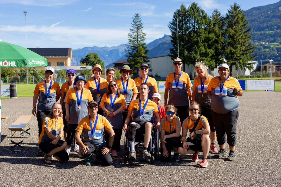 Eine Gruppe von Menschen mit orangefarbenen Hemden und Medaillen posiert an einem sonnigen Tag im Freien mit Bergen, Bäumen und Gebäuden im Hintergrund. Einige Personen stehen, andere knien, und eine Person sitzt im Rollstuhl.