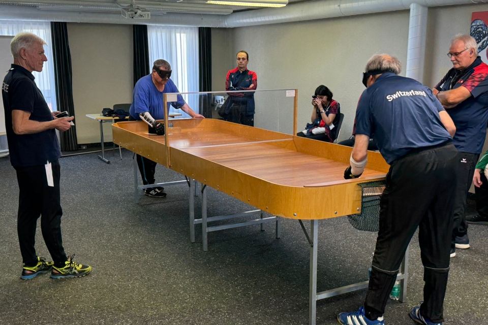 Vier Männer spielen eine Partie Showdown, eine Sportart für Sehbehinderte, an einem Holztisch mit erhöhten Seitenwänden und einer durchsichtigen Trennwand, während zwei Personen im Hintergrund zusehen.