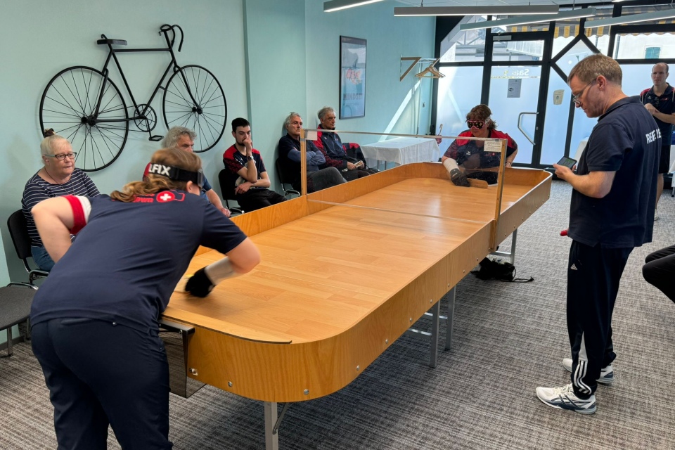 Zwei Personen spielen eine Partie Showdown an einem Holztisch, während mehrere Zuschauer zusehen. Der Raum hat hellblaue Wände, eine Glastür und ein dekoratives Fahrrad an der Wand.