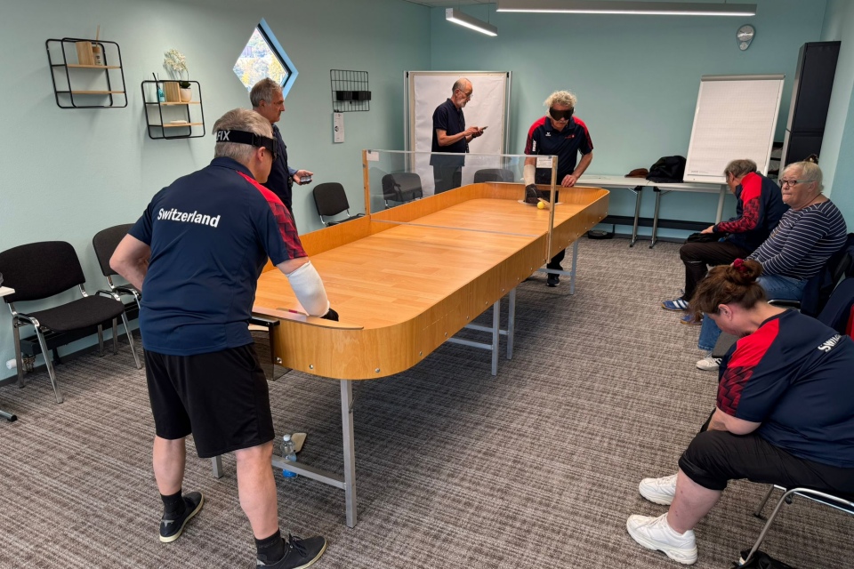 Mehrere Personen in Trikots der Schweizer Mannschaft sind um einen rechteckigen Tisch mit einer durchsichtigen Trennwand versammelt und scheinen in einem hell erleuchteten Raum mit Stühlen und einem Flipchart ein Spiel zu spielen oder zu beobachten.