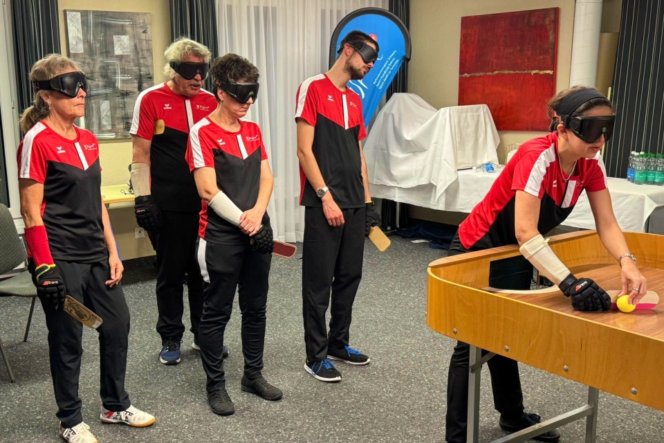 Fünf Personen mit Augenbinden und passenden Uniformen stehen um einen Tisch mit Schlägern, während sich eine Person darauf vorbereitet, einen Ball zu spielen. Im Hintergrund sind Wasserflaschen und abgedeckte Gegenstände zu sehen.