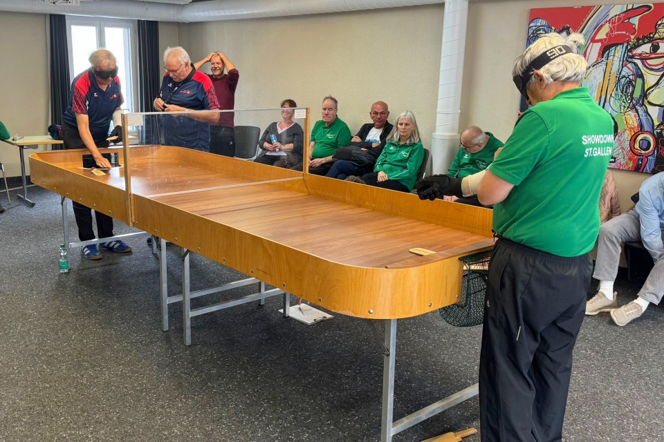 Eine Gruppe von Personen sieht zwei Männern zu, die an einem langen Holztisch ein Spiel spielen; ein Spieler trägt ein grünes Hemd und einen Augenschutz, während das Publikum an der Wand sitzt. Der Schauplatz scheint eine Freizeitveranstaltung in einer Halle zu sein.