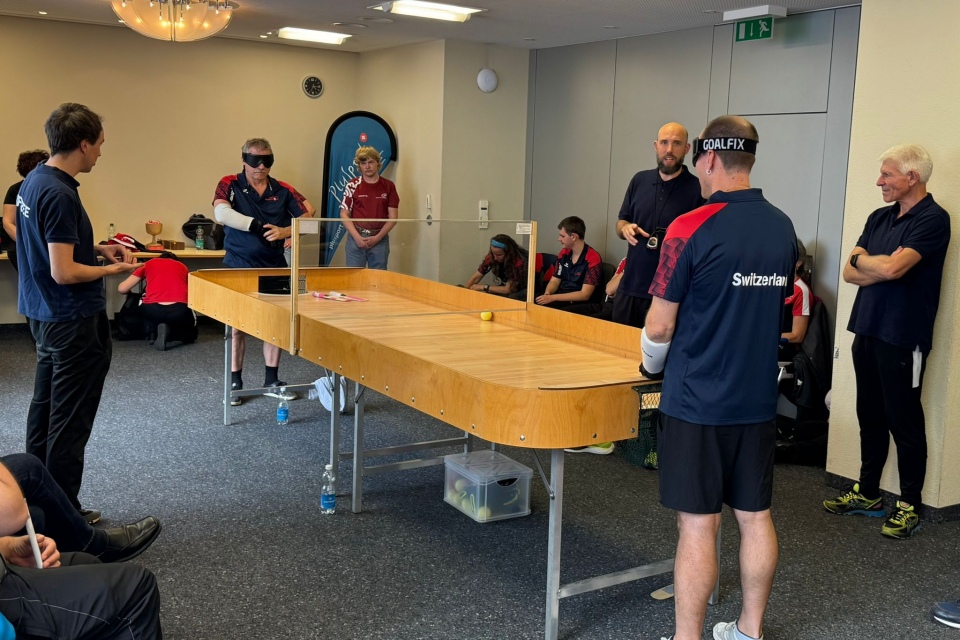 Mehrere Personen spielen und beobachten in einem Raum eine Partie Showdown, eine Sportart für Sehbehinderte. Die Spieler tragen eine Sonnenbrille und schlagen mit Schlägern einen Ball über einen Holztisch mit hohen Seiten. Die Zuschauer stehen und sitzen in der Nähe.