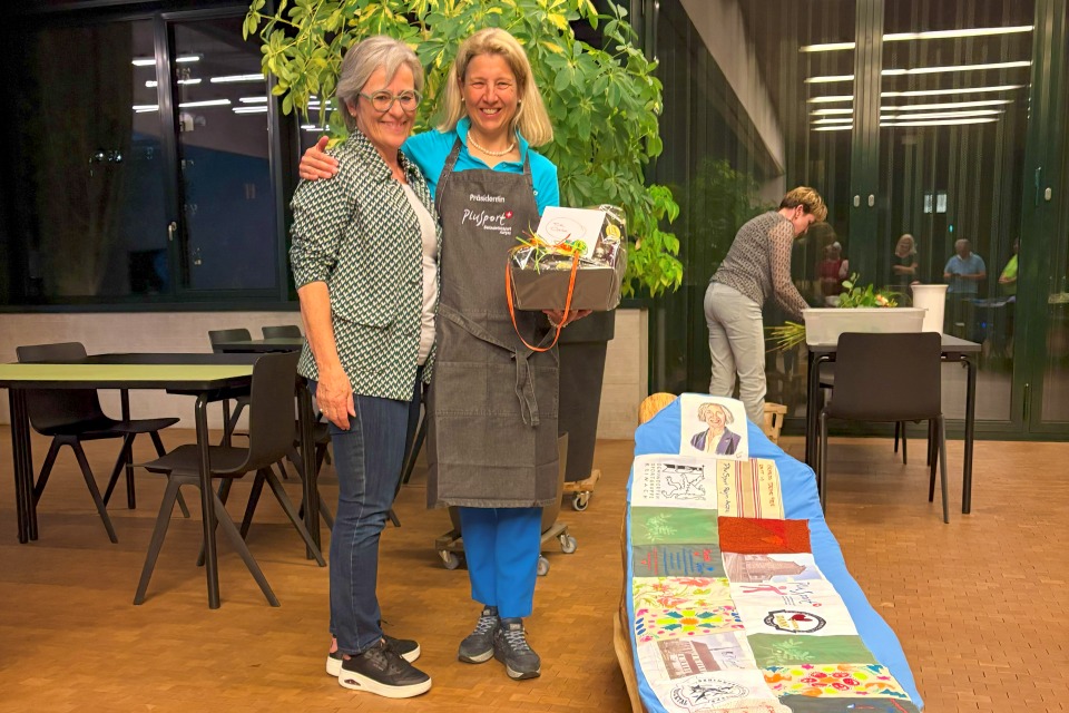 Zwei lächelnde Frauen stehen im Innenbereich zusammen. Eine hält einen Geschenkkorb und trägt eine Schürze. Zu ihren Füßen hängt ein buntes, verziertes Stoffbanner. Im Hintergrund arbeiten andere Personen an Tischen in der Nähe einer großen Fabrik.