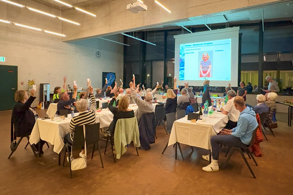Eine Gruppe von Personen sitzt in einem gut beleuchteten Raum an Tischen und hebt während einer Sitzung oder Veranstaltung ihre Hand zur Stimmabgabe. Eine Präsentation mit Foto und Text wird auf eine Leinwand an der Vorderseite des Raumes projiziert.
