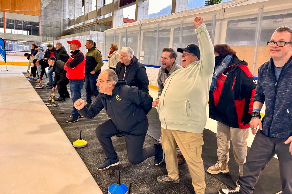 Eine Gruppe von Menschen jubelt begeistert am Rande einer Eishalle, einige knien nieder und heben begeistert die Fäuste. Sie beobachten eine Veranstaltung und scheinen sich gemeinsam zu amüsieren.