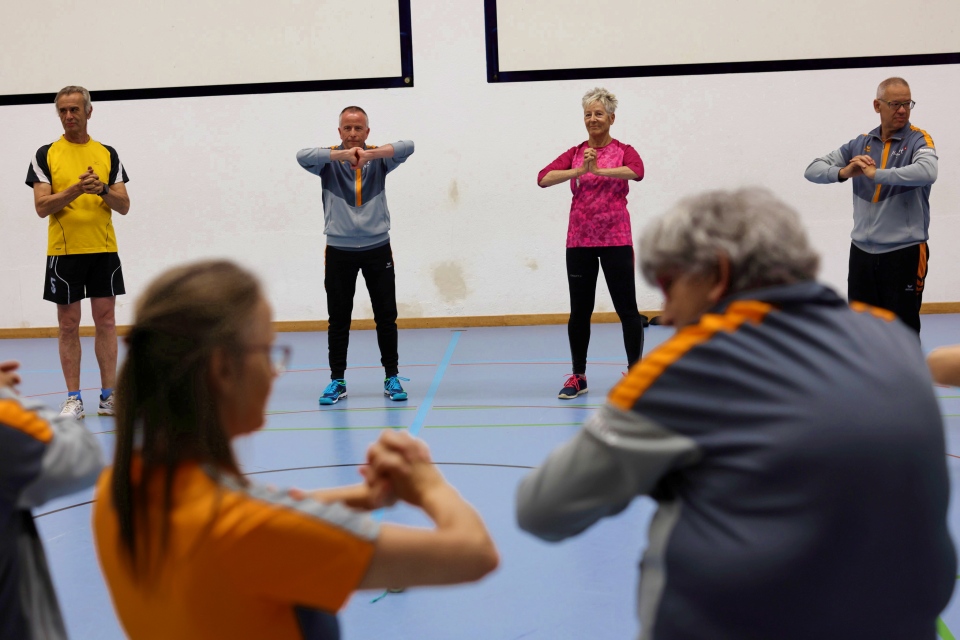 Eine Gruppe Erwachsener steht in einer Turnhalle im Halbkreis und macht Armübungen. Einige tragen passende Sportuniformen, während eine Frau in Pink hervorsticht. Sie scheinen an einem Gruppenfitnesskurs teilzunehmen.