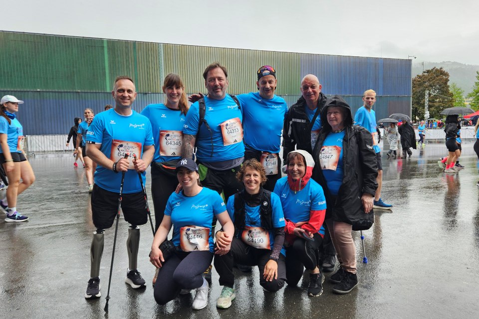 Eine Gruppe von neun Personen in blauen Hemden, darunter zwei mit Beinprothesen, posiert und lächelt gemeinsam an einem nassen, regnerischen Tag bei einer Laufveranstaltung im Freien. Im Hintergrund sind andere Teilnehmer und ein großes Industriegebäude zu sehen.