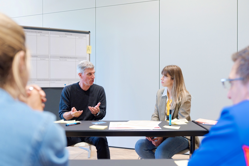 Vier Personen sitzen in einem hellen, modernen Büro um einen Tisch und diskutieren. Ein Mann gestikuliert mit den Händen, während er mit einer Frau spricht. Im Hintergrund ist ein Flipchart mit Notizen zu sehen.
