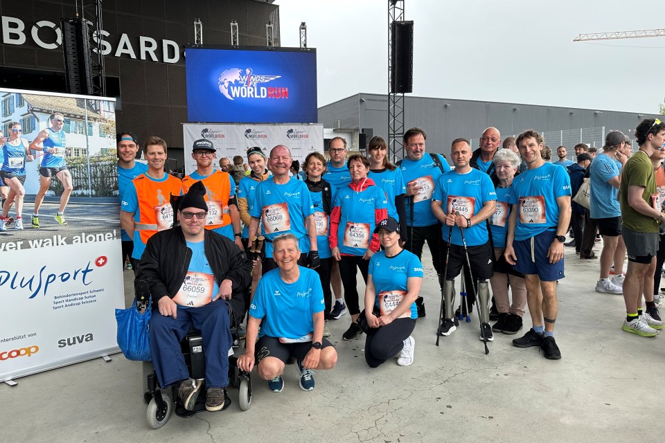 Eine Gruppe von Menschen in blauen Hemden, von denen einige eine Behinderung haben, posieren zusammen und lächeln für ein Foto bei einer Laufveranstaltung im Freien. Auf einem großen Schild und einem Bildschirm hinter ihnen steht World Run. Im Hintergrund sind weitere Teilnehmer zu sehen.