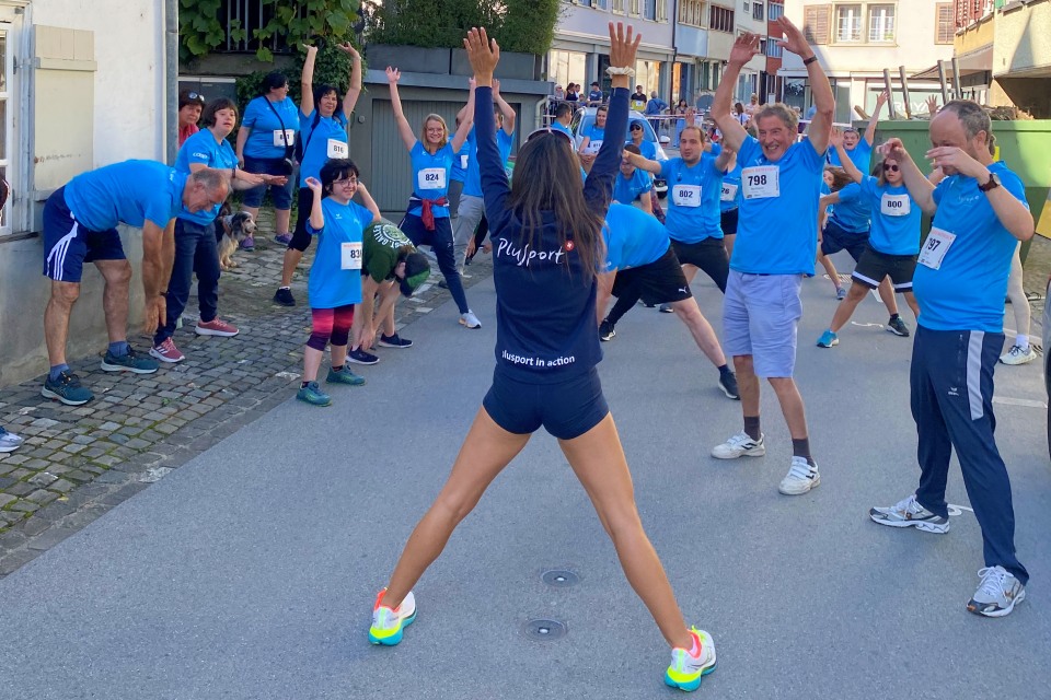 Eine Gruppe von Menschen in blauen Hemden und Startnummern dehnt und trainiert gemeinsam im Freien, angeführt von einer Frau in Sportkleidung, die ihnen mit erhobenen Armen auf einer Straße in einem Wohngebiet gegenübersteht.