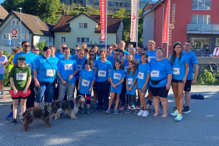 Eine Gruppe von Menschen in blauen Hemden und mit Startnummern posiert an einem sonnigen Tag im Freien vor Häusern und Gebäuden; in der Gruppe sind auch zwei Hunde zu sehen.