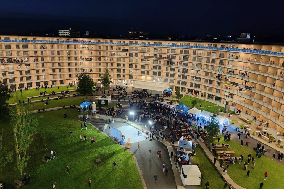 Eine nächtliche Veranstaltung unter freiem Himmel, bei der sich viele Menschen in einem grasbewachsenen Innenhof versammeln, der von einem großen, geschwungenen Wohngebäude mit vielen beleuchteten Fenstern und Balkonen umgeben ist. Auf dem Rasen sind Zelte und Picknicktische aufgestellt.