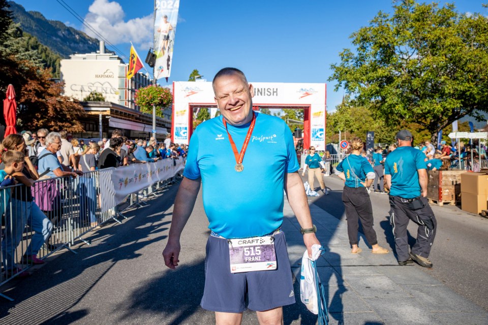 Ein lächelnder Mann in blauem Hemd, Shorts und Startnummer steht an der Ziellinie eines Marathons, hält eine Tasche in der Hand und zeigt seine Medaille, während im Hintergrund Zuschauer und andere Teilnehmer zu sehen sind.