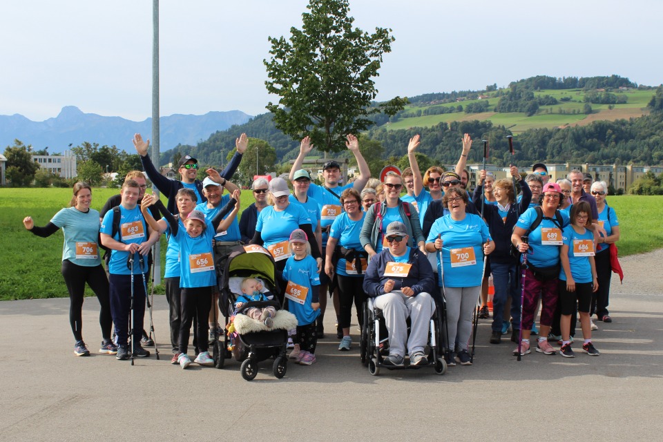 Eine große Gruppe von Menschen in blauen Hemden, einige mit nummerierten Lätzchen, lächeln und winken im Freien. Einige benutzen Gehstöcke, eine Person sitzt im Rollstuhl und ein Kind ist in einem Kinderwagen unterwegs. Im Hintergrund sind grüne Hügel und Bäume zu sehen.