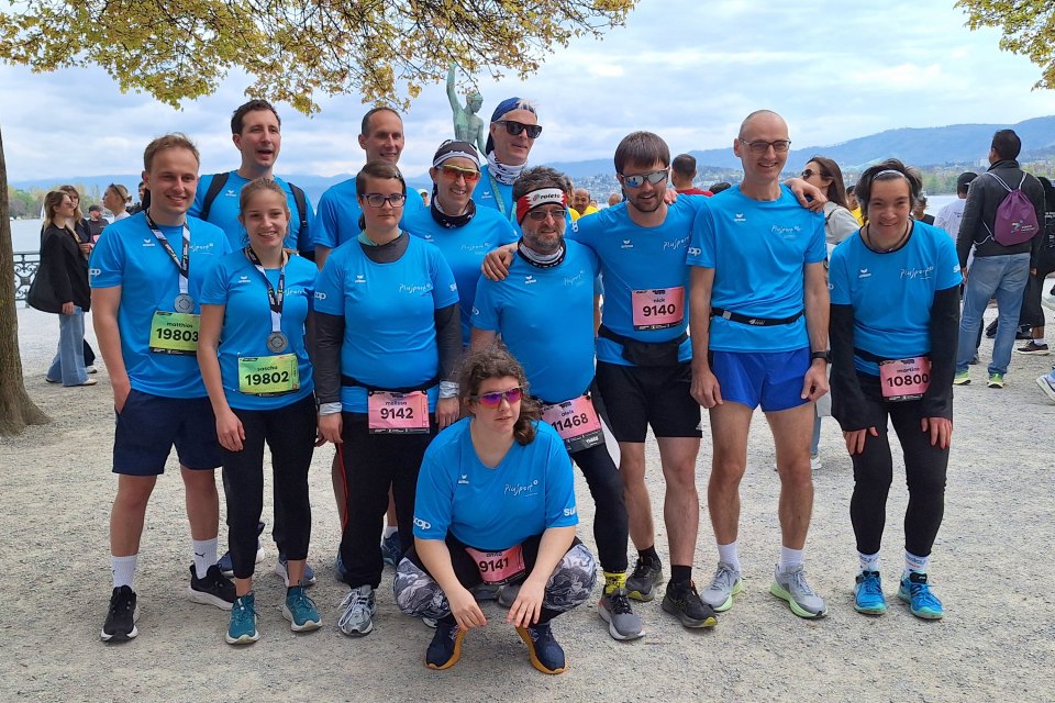 Eine Gruppe von dreizehn Personen in passenden blauen Laufshirts posiert nach einem Rennen im Freien. Einige tragen Medaillen und nummerierte Startnummern. Im Hintergrund sind Bäume und andere Menschen zu sehen.