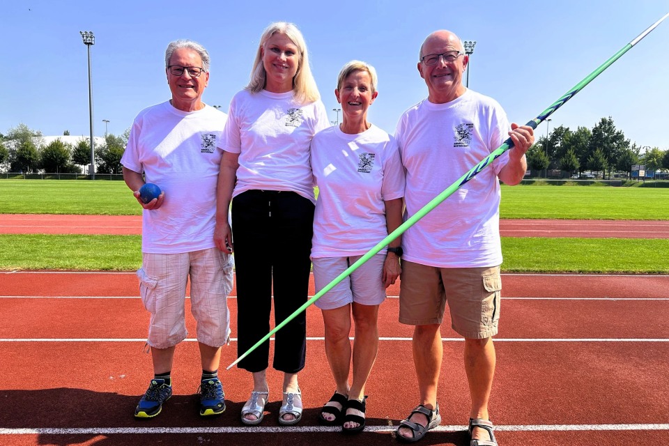 Vier ältere Erwachsene stehen nebeneinander auf einem Leichtathletikfeld, alle tragen passende weiße T-Shirts. Zwei von ihnen halten ein Sportgerät - ein Kugelstoßgerät und einen Speer. Sie lächeln und posieren für das Foto an einem sonnigen Tag.