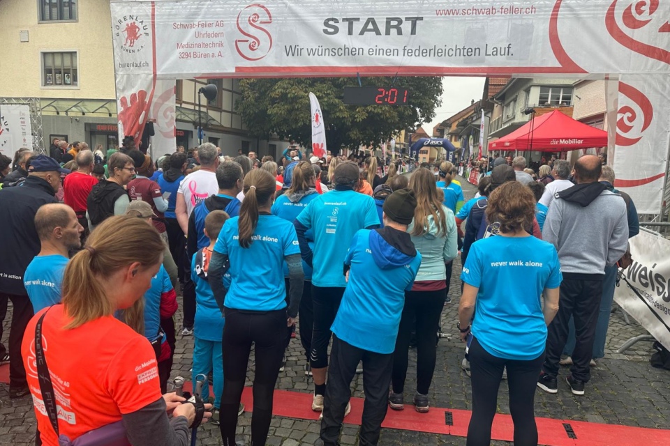 Eine Schar von Läufern, viele in blauen Hemden, versammelt sich an der Startlinie einer Laufveranstaltung auf einer Kopfsteinpflasterstraße unter einem Banner mit der Aufschrift "Start" auf Deutsch. Der Timer über dem Banner zeigt 2:01 an.
