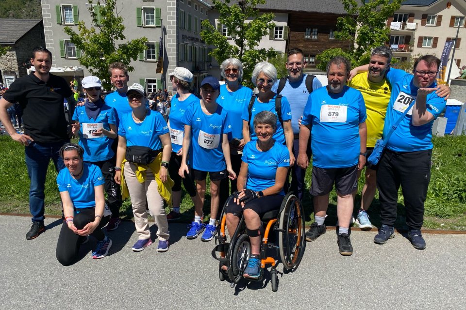 Eine Gruppe lächelnder Läufer, die meisten in blauen Hemden und Startnummern, posiert an einem sonnigen Tag im Freien. Ein Teilnehmer sitzt in einem Rollstuhl. Im Hintergrund sind grüne Bäume und Gebäude zu sehen.