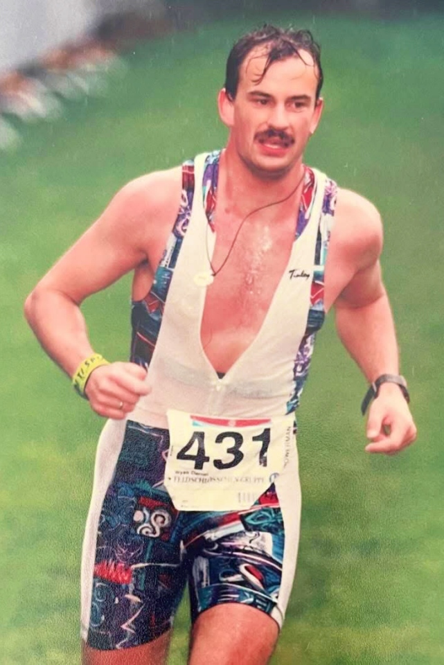 Ein Mann mit Schnurrbart läuft im Freien und trägt einen bunten Triathlonanzug mit der Nummer 431. Er schwitzt und scheint konzentriert zu sein. Er trägt eine Uhr am Handgelenk und ein Armband am Arm und rennt auf einer grasbewachsenen Fläche.