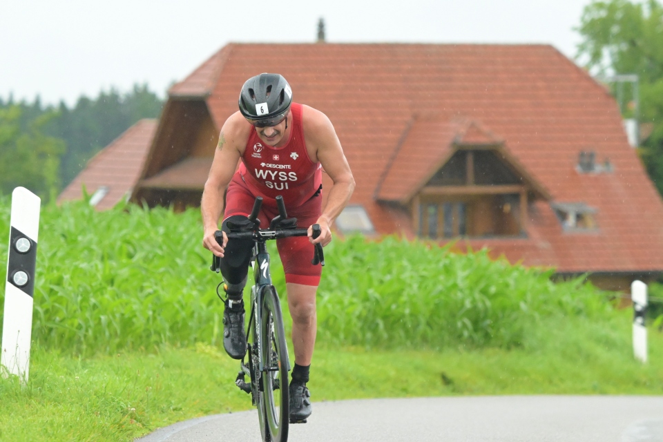 Ein Triathlet in rotem Anzug und schwarzem Helm fährt mit dem Fahrrad auf einer Landstraße mit grünem Gras und einem Haus mit rotem Dach im Hintergrund. Der Sportler wirkt konzentriert und lehnt sich auf dem Fahrrad nach vorne.