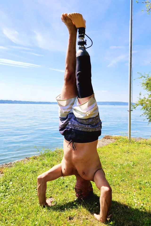 Ein Mann ohne Hemd, in Badehose und mit Beinprothese, balanciert im Kopfstand auf einer Wiese an einem See, mit blauem Himmel und ruhigem Wasser im Hintergrund.