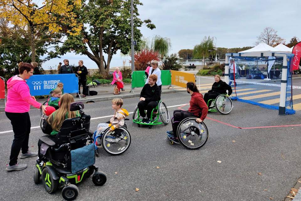 Menschen verschiedenen Alters, einige in Rollstühlen, spielen auf einem gepflasterten Platz im Freien ein Spiel. Im Hintergrund sind bunte Absperrungen, Bäume mit Herbstlaub und Zelte zu sehen. Auf der linken Seite steht ein Trainer, der die Teilnehmer anleitet.